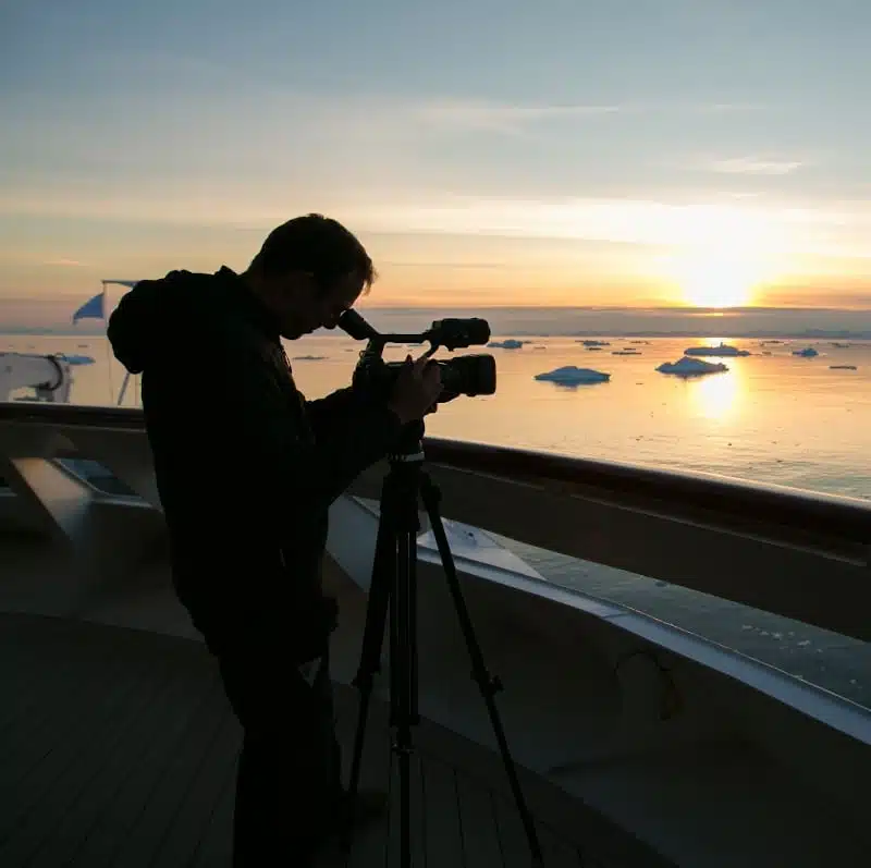 Silhouette d’un vidéaste filmant un coucher de soleil sur une mer parsemée d’icebergs.