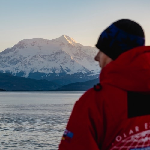 Passager en parka regardant vers la mer surmontée d'une montagne enneigée
