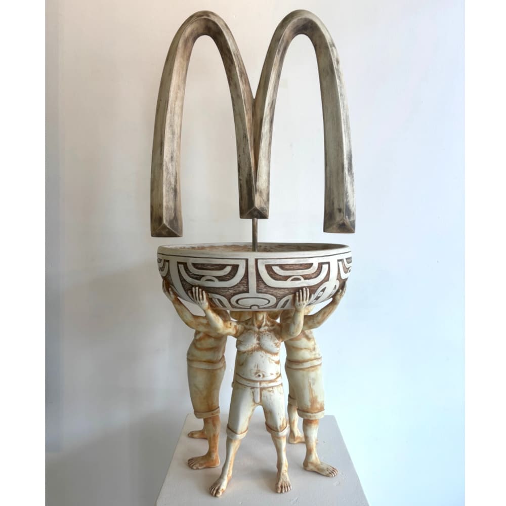 A sculpture by Tahe: three people holding up a traditional bowl with a large wooden McDonald’s “M” on it