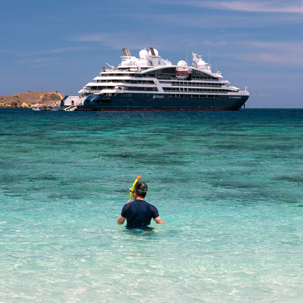 A PONANT passenger by the water’s edge with a mask and snorkel, and the PONANT EXPLORATIONS vessel on the horizon